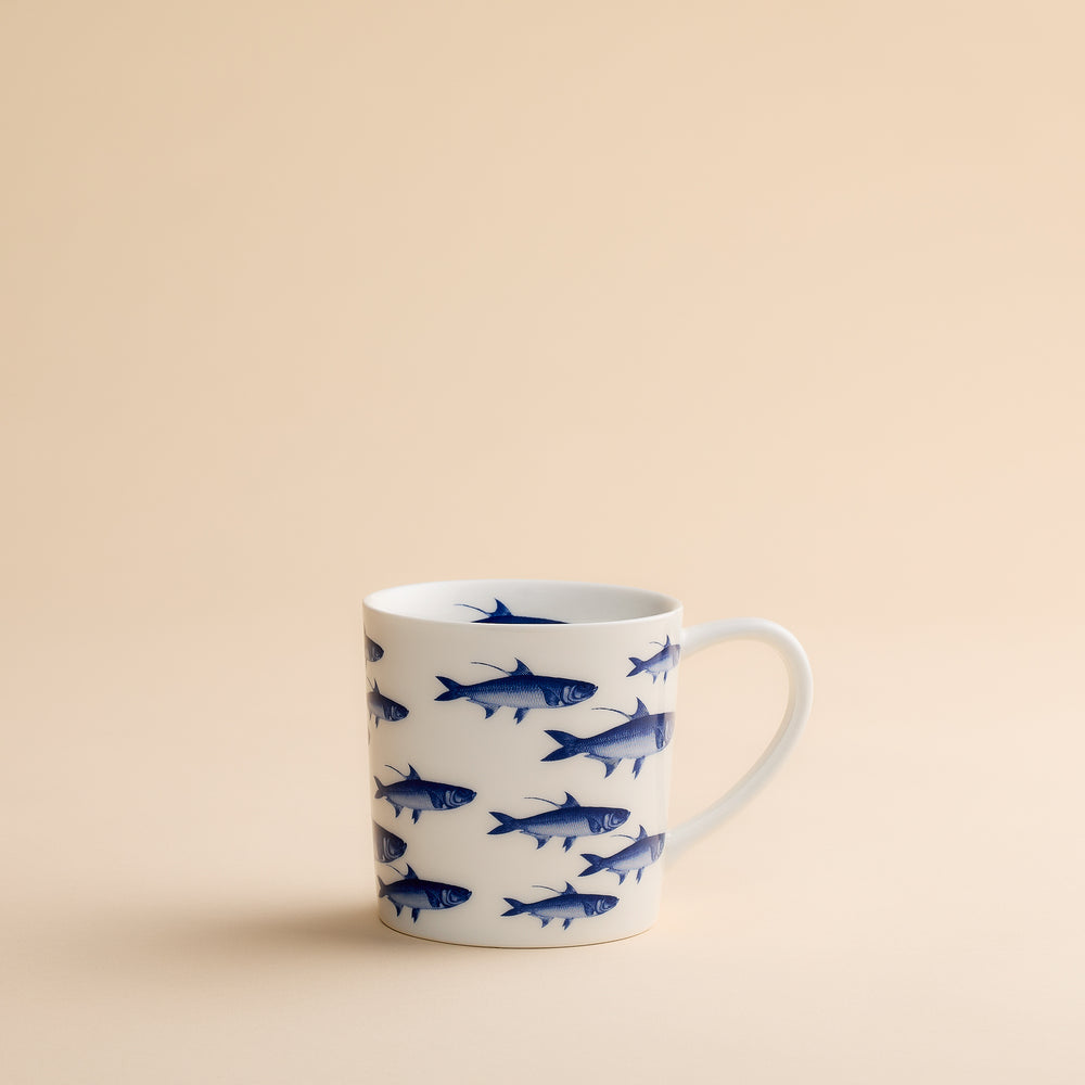 The School of Fish Mug by Caskata Artisanal Home features blue fish illustrations on a white ceramic surface and is displayed against a plain beige background. Made from high-fired porcelain, this coastal mug ensures both durability and enduring charm.