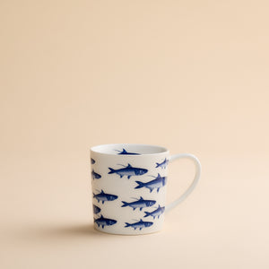 The School of Fish Mug by Caskata Artisanal Home features blue fish illustrations on a white ceramic surface and is displayed against a plain beige background. Made from high-fired porcelain, this coastal mug ensures both durability and enduring charm.