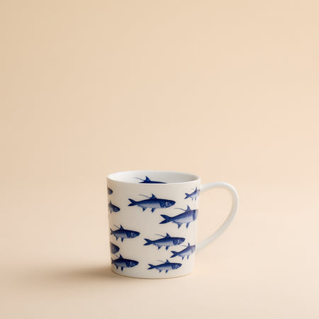 The School of Fish Mug by Caskata Artisanal Home features blue fish illustrations on a white ceramic surface and is displayed against a plain beige background. Made from high-fired porcelain, this coastal mug ensures both durability and enduring charm.