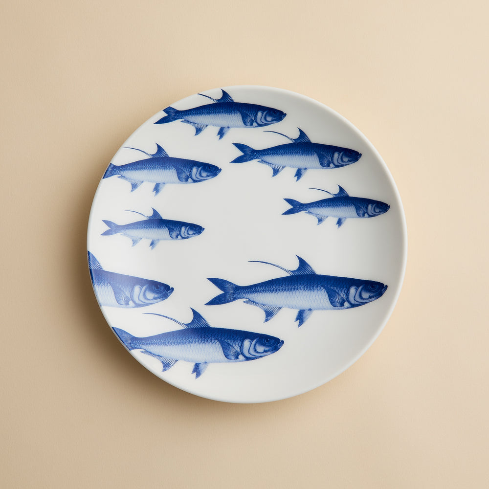 The School of Fish Coupe Salad Plate by Caskata Artisanal Home, a white porcelain plate adorned with charming blue fish illustrations, is displayed on a beige background.
