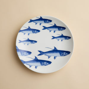 The School of Fish Coupe Salad Plate by Caskata Artisanal Home, a white porcelain plate adorned with charming blue fish illustrations, is displayed on a beige background.