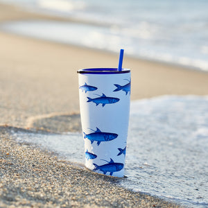 The Caskata School of Fish Stainless Steel Insulated Tumbler, featuring blue fish illustrations and a blue straw, rests on wet sand by the shore with gentle ocean waves in the background.