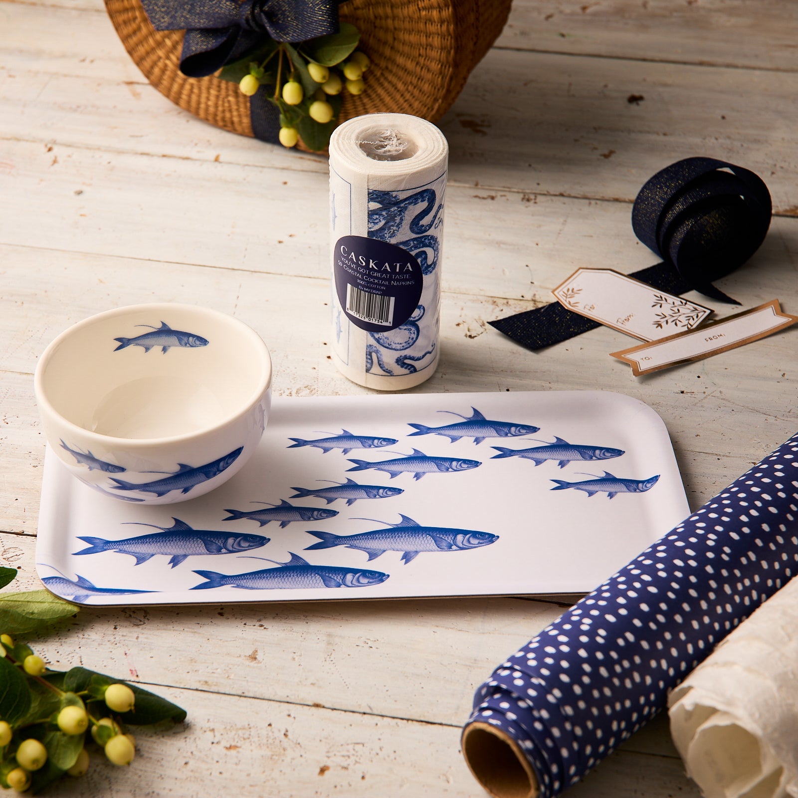 The Caskata "Everything's Going Swimmingly" Bundle, featuring a blue School of Fish appetizer tray and bowl set, is displayed on a wooden table with wrapping paper, ribbon, cocktail napkins, gift tags, flowers, and a wicker basket nearby.
