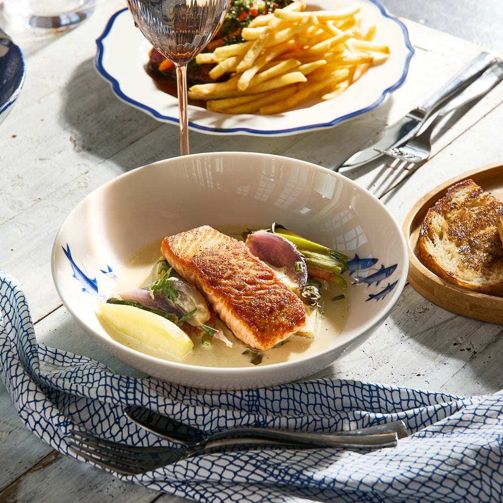 A bowl featuring seared salmon, vegetables, and sauce rests on the table next to a plate of fries, a glass of wine, and sliced bread. The Caskata Coastal Icons Entrée Bowls, Mixed Set of 4, from our coastal dinnerware collection perfectly complement the fresh meal and are microwave safe for added convenience.