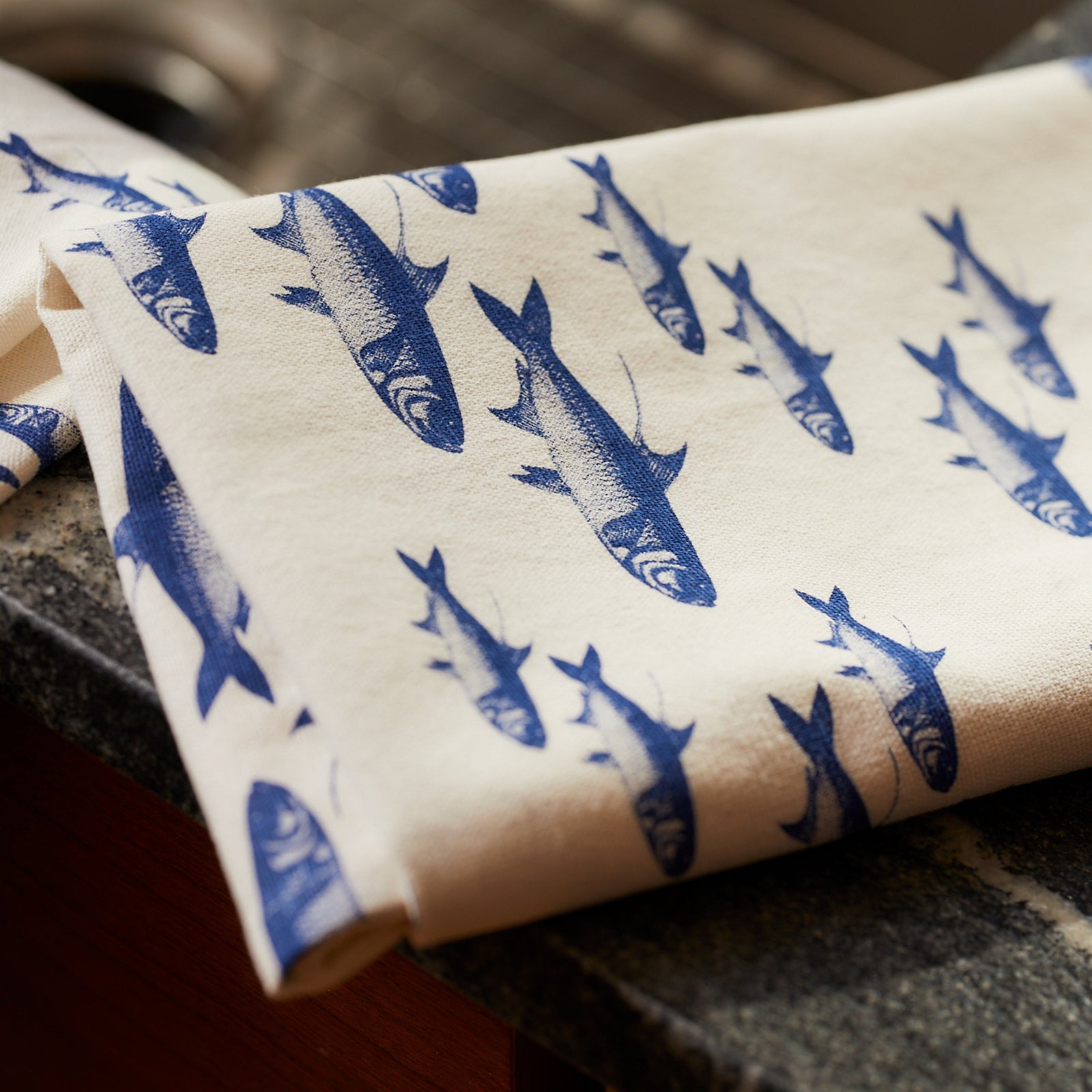 The "Fishing Fun" Bundle by Caskata, featuring a blue School of Fish porcelain platter and a matching kitchen towel, is displayed on a wooden table with ribbons, a woven basket, and rolled white fabric.