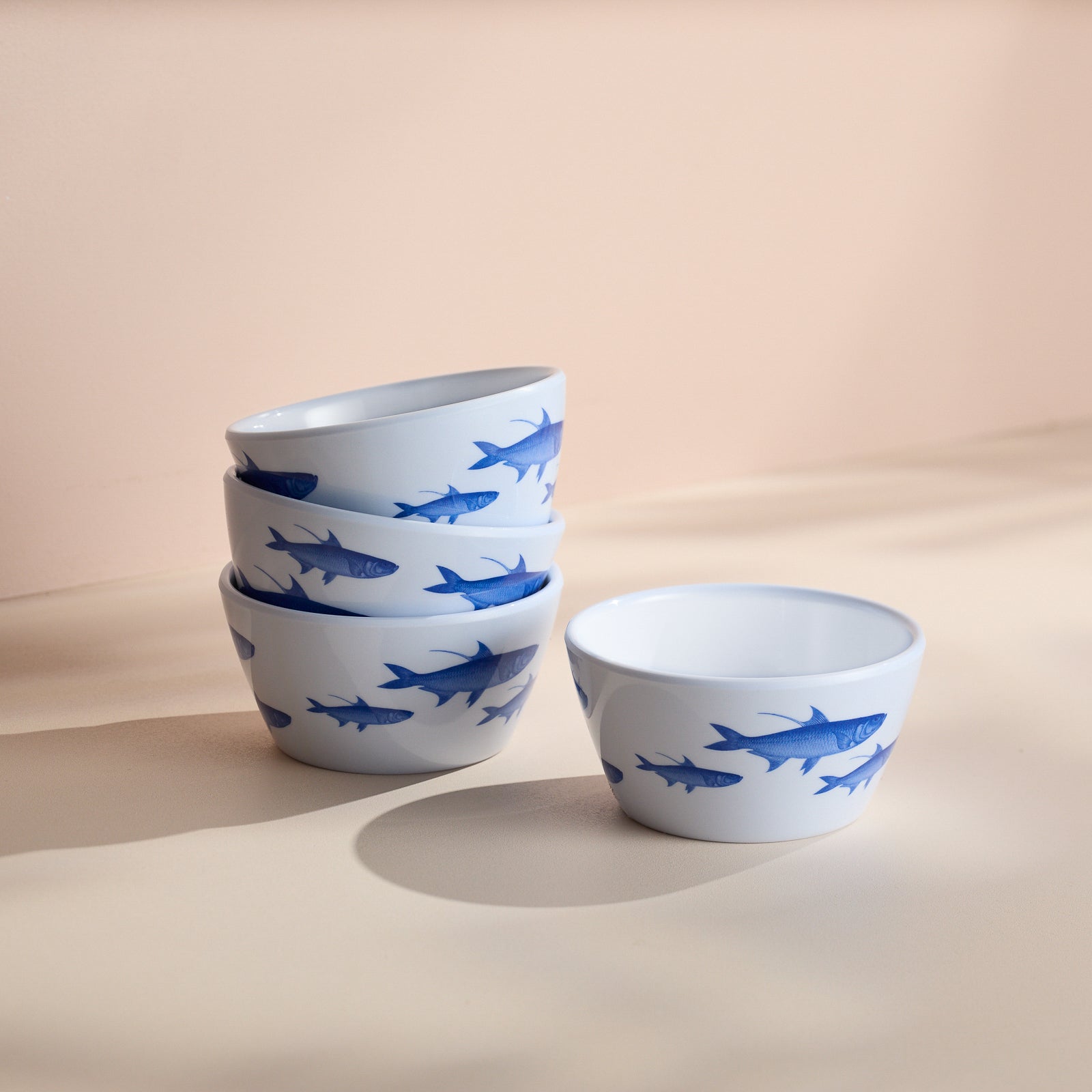 The "School of Fish Melamine Snack Bowls, Set of 4" by Caskata features four stacked white bowls adorned with blue fish designs, alongside a matching bowl on a light surface. These outdoor dinnerware pieces are crafted from BPA-free melamine for safe and stylish dining.