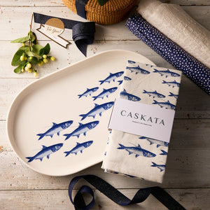 The Caskata Fishing Fun Bundle, including a School of Fish porcelain platter and matching kitchen towel, rests on a rustic wooden table beside gift wrap, ribbon, and a decorative branch.