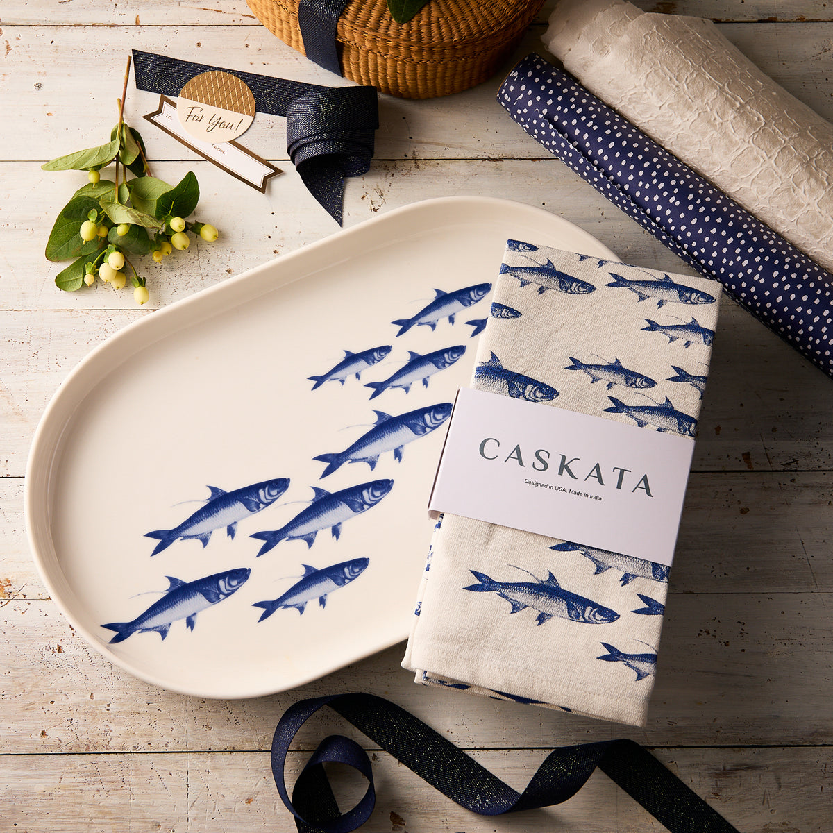 The "Fishing Fun" Bundle by Caskata, featuring a blue School of Fish porcelain platter and a matching kitchen towel, is displayed on a wooden table with ribbons, a woven basket, and rolled white fabric.