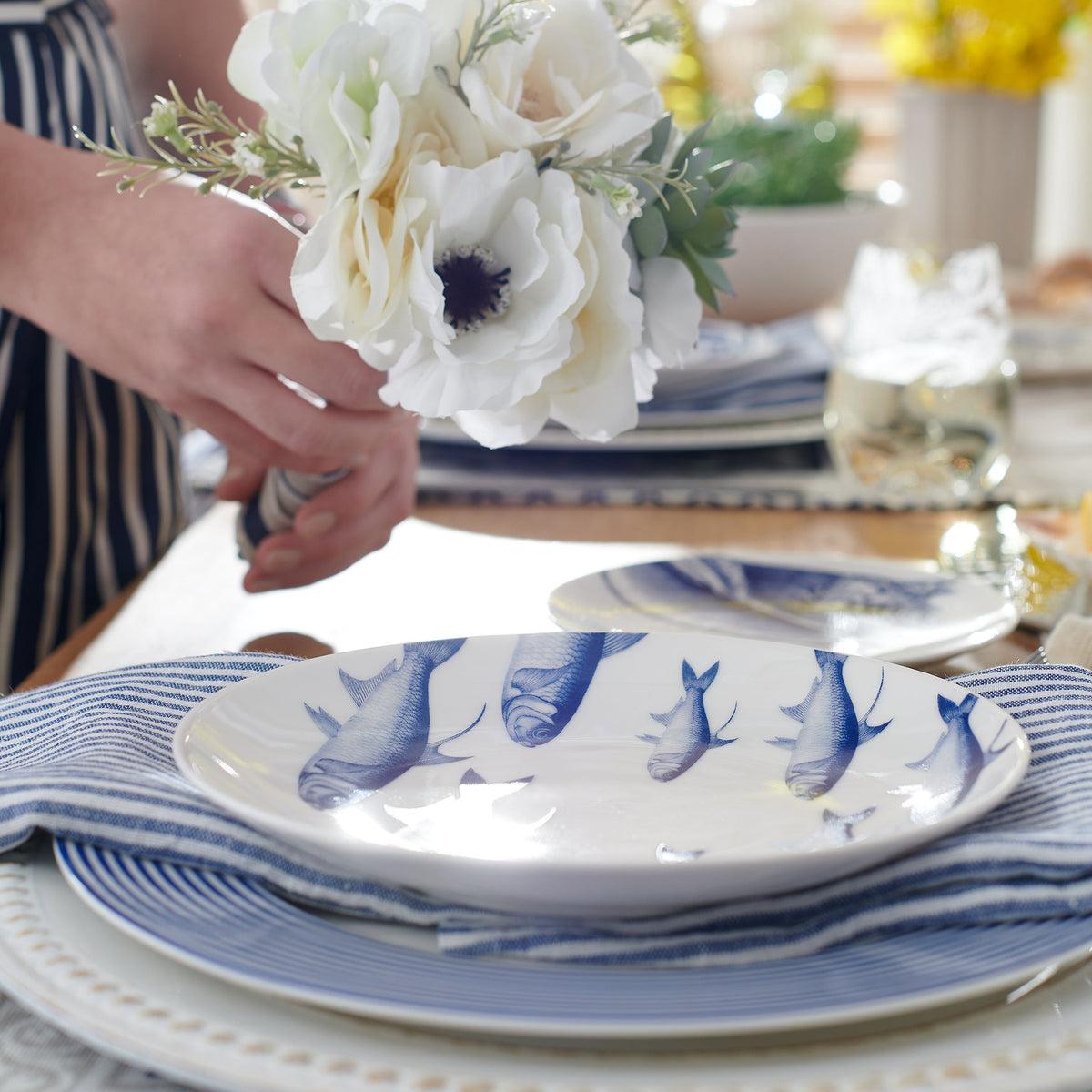 A woman is setting a table with blue and white School of Fish Coupe Salad Plates adorned with accent plates and flowers from Caskata Artisanal Home.