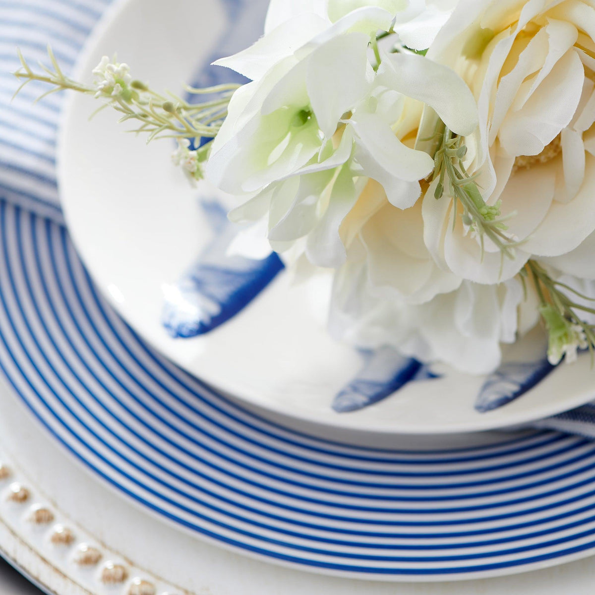 Close-up of a Caskata Artisanal Home Newport Stripe Rimmed Dinner Plate with blue stripes, white flowers, and a striped napkin. Another plate featuring elegant blue designs is partially visible underneath, showcasing the beauty of contemporary dinnerware.