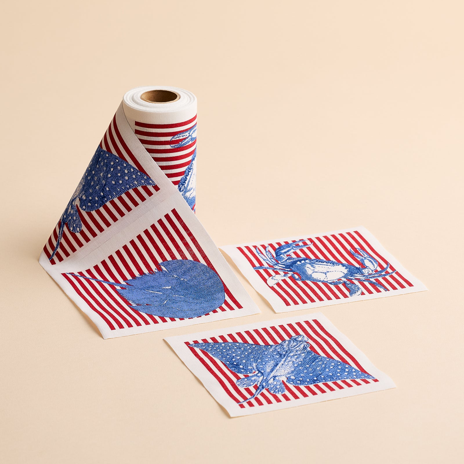 The Seaside Stripes Cocktail Napkin Roll by MY DRAP features a roll of patterned napkins adorned with red stripes alongside blue eagle, heart, and deer motifs, reminiscent of coastal charm; two individual sheets are displayed flat on a beige background.