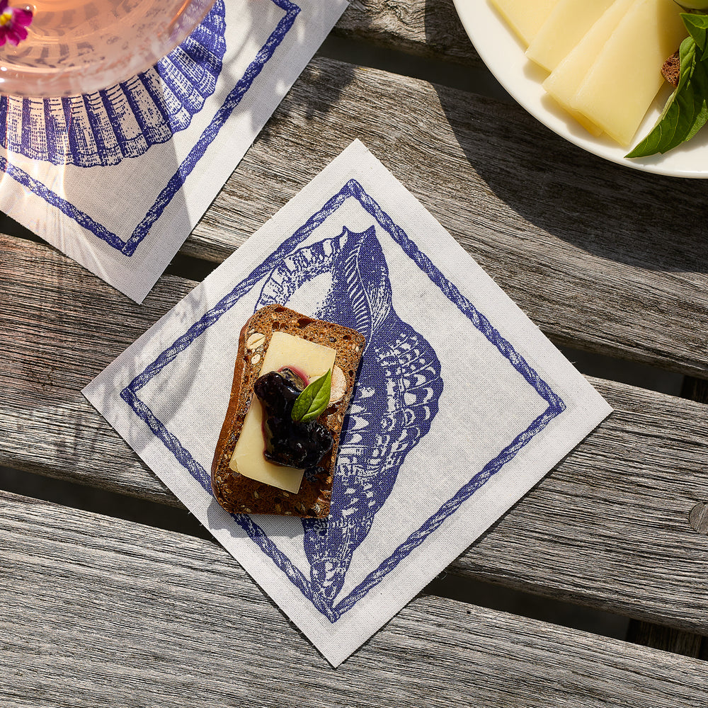 A slice of bread with cheese, jam, and basil rests on a blue and white Shells Cocktail Napkin Roll by MY DRAP—crafted from 100% cotton napkins—making for a stylish party essential atop a wooden surface.