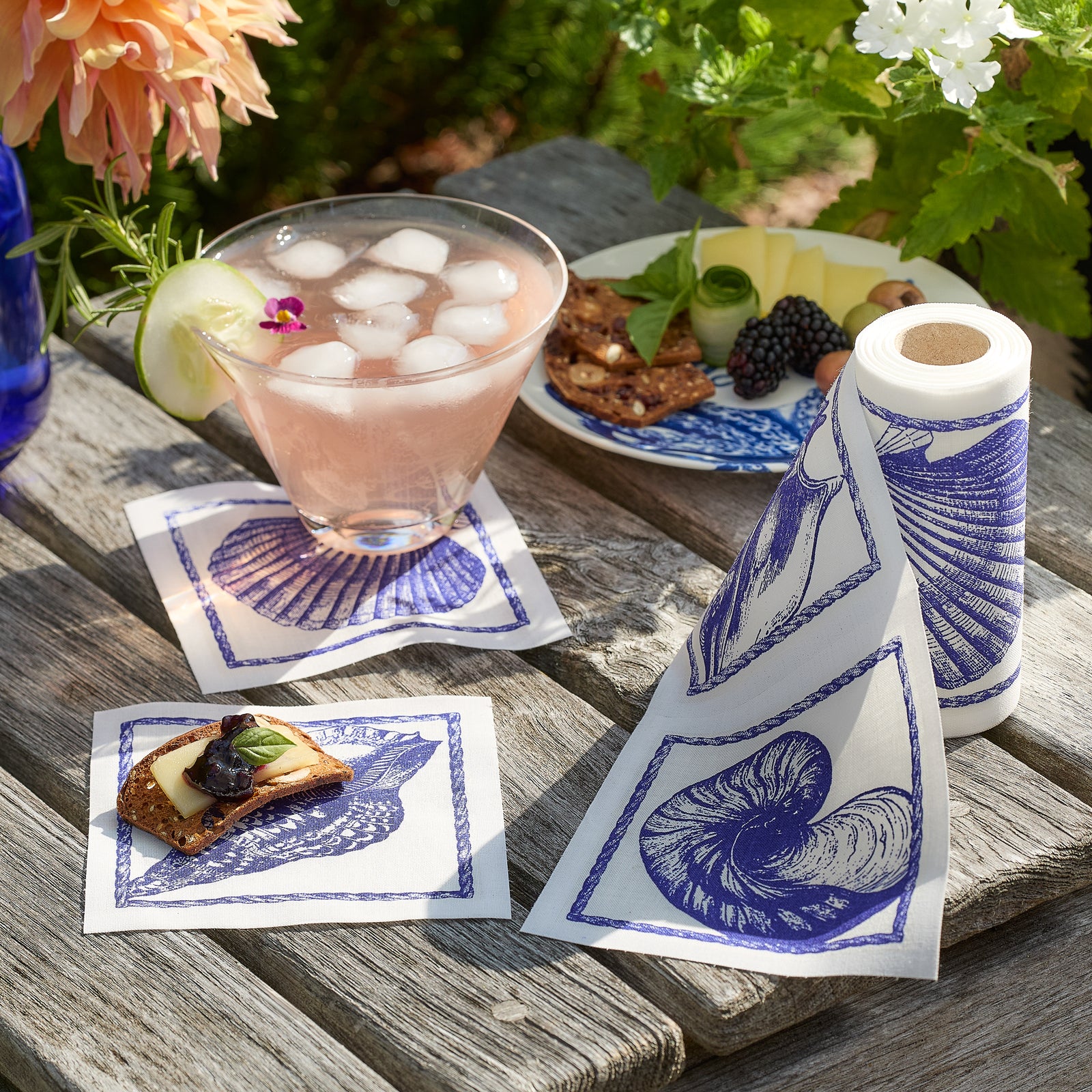 A roll of MY DRAP Shells Cocktail Napkin Roll, decorated with blue seashell and marine designs, is displayed with three square napkins cut and arranged beside it on a beige background—these napkins are a must-have party essential for any celebration.