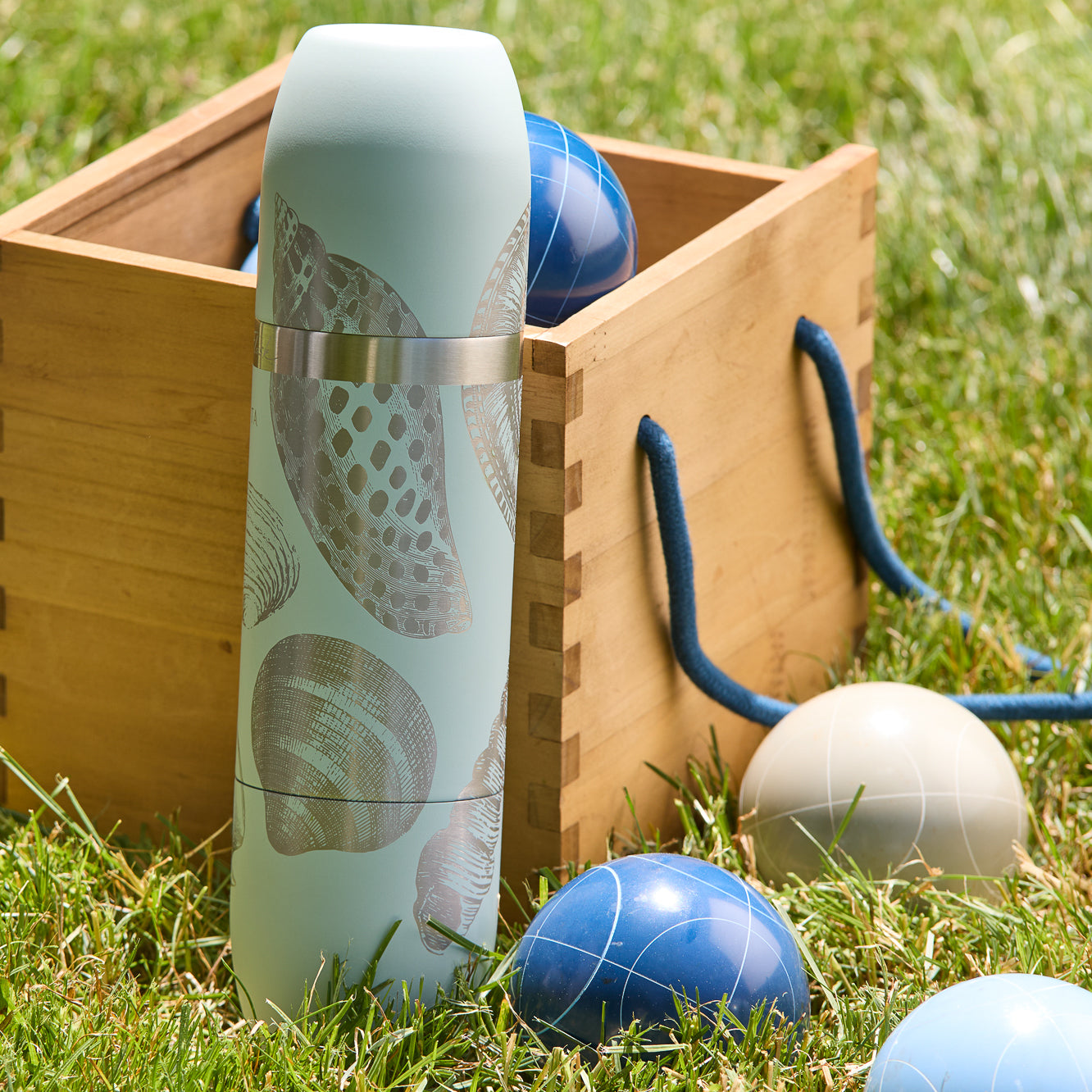 The Caskata x High Camp Light Blue Shells Wine Flask, a cylindrical outdoor wine flask with seashell illustrations, is shown by a Caskata box labeled "Parkside Flask" and "All-in-one Insulated Wine Set," featuring magnetic cups for easy sipping.