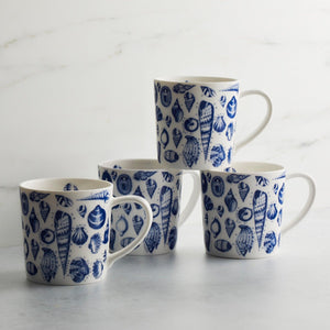Four Shells Mugs from Caskata Artisanal Home made of high-fired porcelain with blue seashell patterns are stacked in a pyramid on a light gray surface against a plain white background.