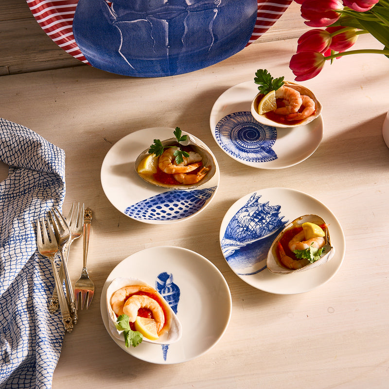 On a wooden table, four Caskata Artisanal Home's Shells Small Plates, adorned with seafood appetizers like shrimp and lemon wedges, are set beside a blue-patterned napkin and silverware. A vase of red tulips adds a touch of color.