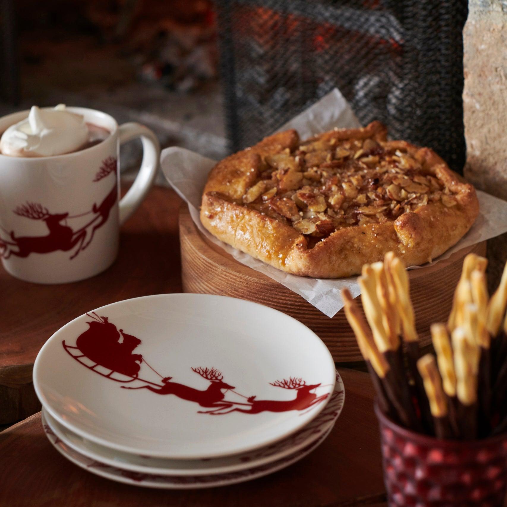 This set of four heirloom-quality Sleigh Small Plates from Caskata Artisanal Home showcases a red design of Santa in a sleigh pulled by reindeer, artfully arranged in an overlapping pattern.