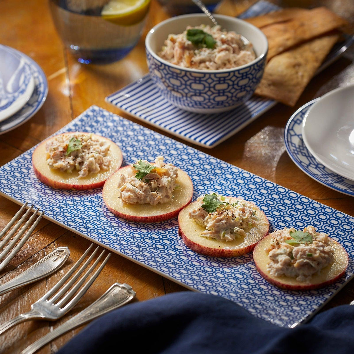 A Newport Petite Serving Tray by Caskata elegantly displays four apple slices topped with chicken salad on a wooden table set with bone china. In the background, a bowl of chicken salad, a glass of lemon water, and utensils complete this charming scene.