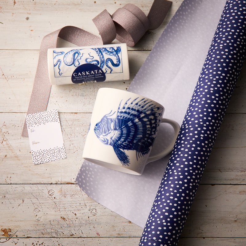 The “Ella in the Morning” Bundle by Caskata features a Spiky Fish Mug with a blue fish design, a Caskata soap bar, polka dot gift tag, ribbon, and blue polka dot wrapping paper arranged on a wooden surface.