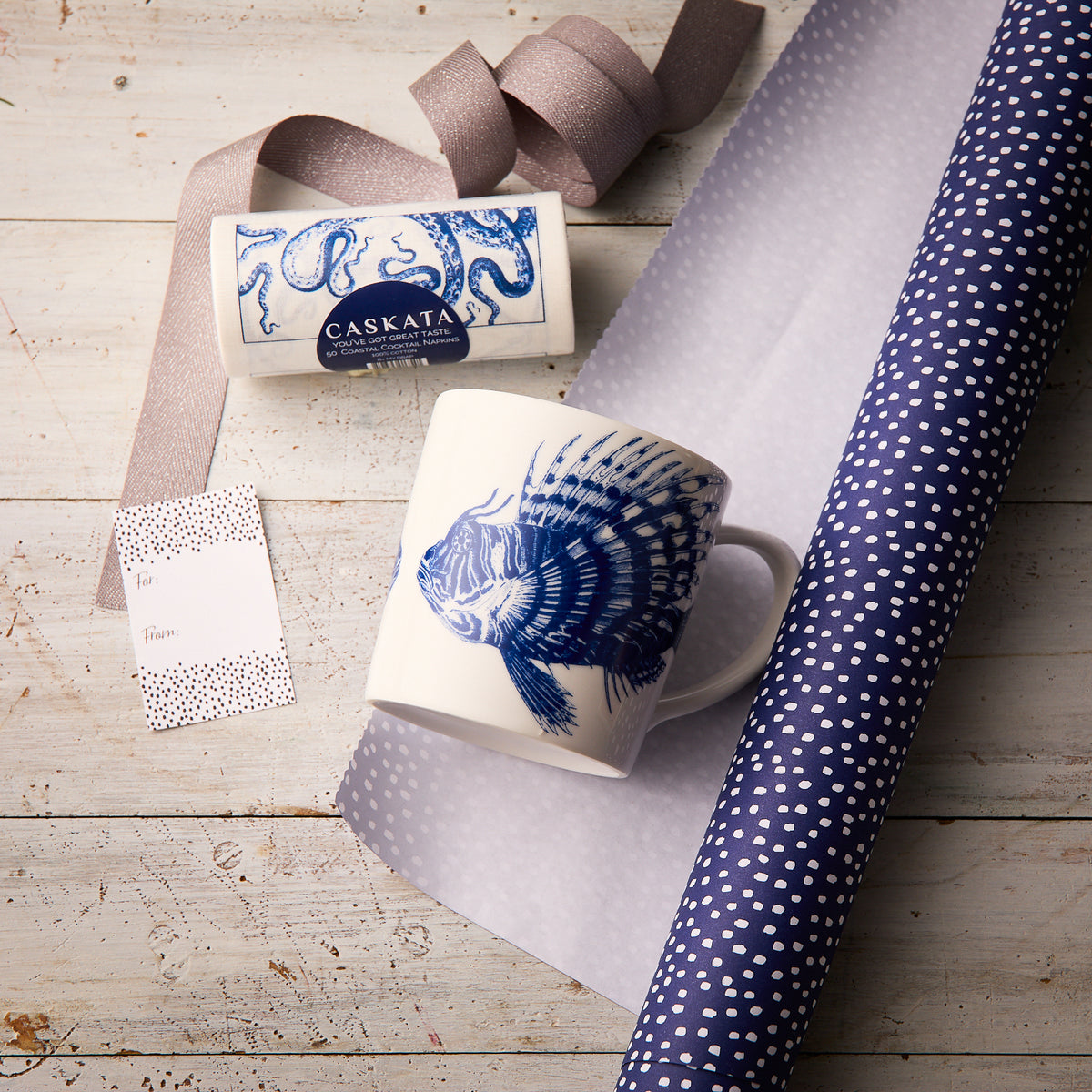 The “Ella in the Morning” Bundle by Caskata features a Spiky Fish Mug with a blue fish design, a Caskata soap bar, polka dot gift tag, ribbon, and blue polka dot wrapping paper arranged on a wooden surface.
