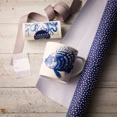 The “Ella in the Morning” Bundle by Caskata features a Spiky Fish Mug with a blue fish design, a Caskata soap bar, polka dot gift tag, ribbon, and blue polka dot wrapping paper arranged on a wooden surface.