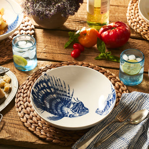 A table set for a meal with Caskata’s Spiky Fish Entrée Bowl featuring a blue fish illustration, blue glasses with ice and lemon, vegetables, bread, wine, a modern serving bowl, and a floral centerpiece on a wooden surface.