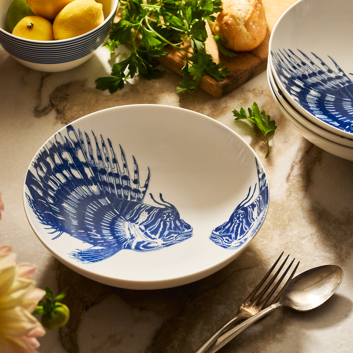 The Spiky Fish Entrée Bowl by Caskata, featuring a blue fish design on white, rests on a marble countertop next to a fork, spoon, parsley, bread, and a bowl of lemons.