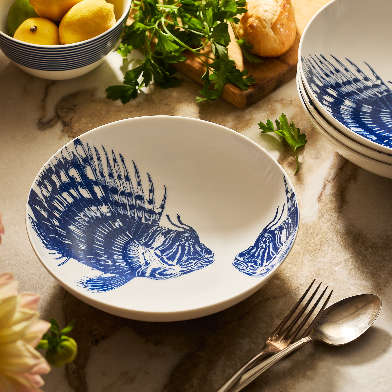 The Caskata Spiky Fish Entrée Bowl is a white shallow bowl with two blue fish illustrations inside, perfect coastal dinnerware for adding a fresh maritime touch.