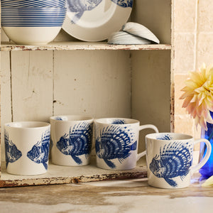 Four Caskata Spiky Fish Mugs with white coastal design and fish illustrations sit on a shelf beside a matching plate and decorative seashells. A flower arrangement is nearby on the counter.