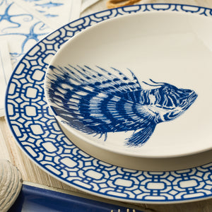 The Caskata Spiky Fish Coupe Salad Plate features a blue fish illustration, making it perfect for coastal dinnerware and pairing beautifully with plates sporting bold geometric blue and white borders.
