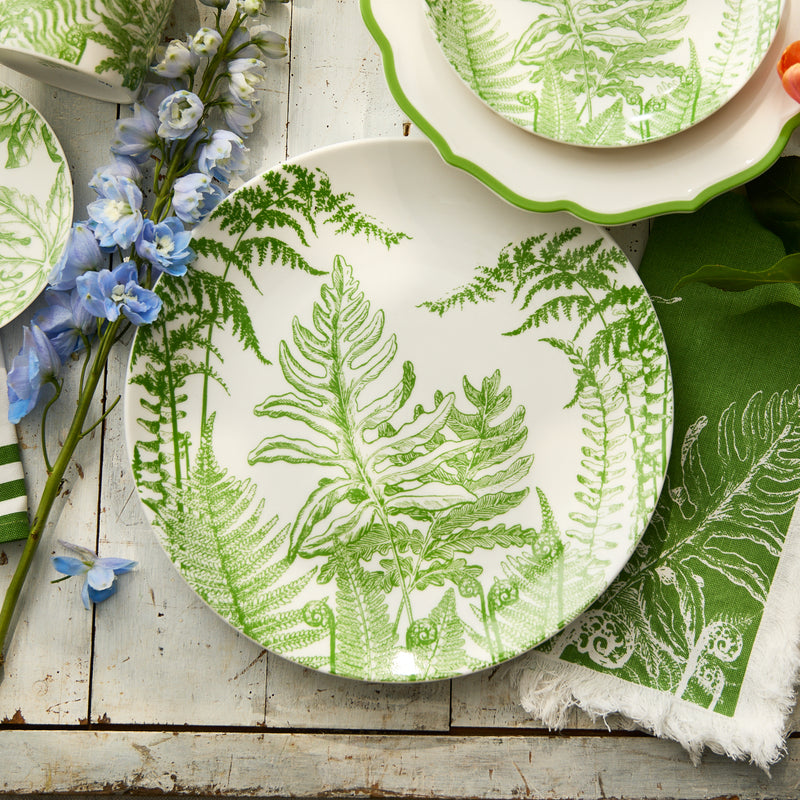 The Caskata Spring Verde Round Platter, premium porcelain with green fern leaf designs, rests beside a blue flower, green napkin, and matching dishware on a rustic wood surface—ideal for stylish serving.