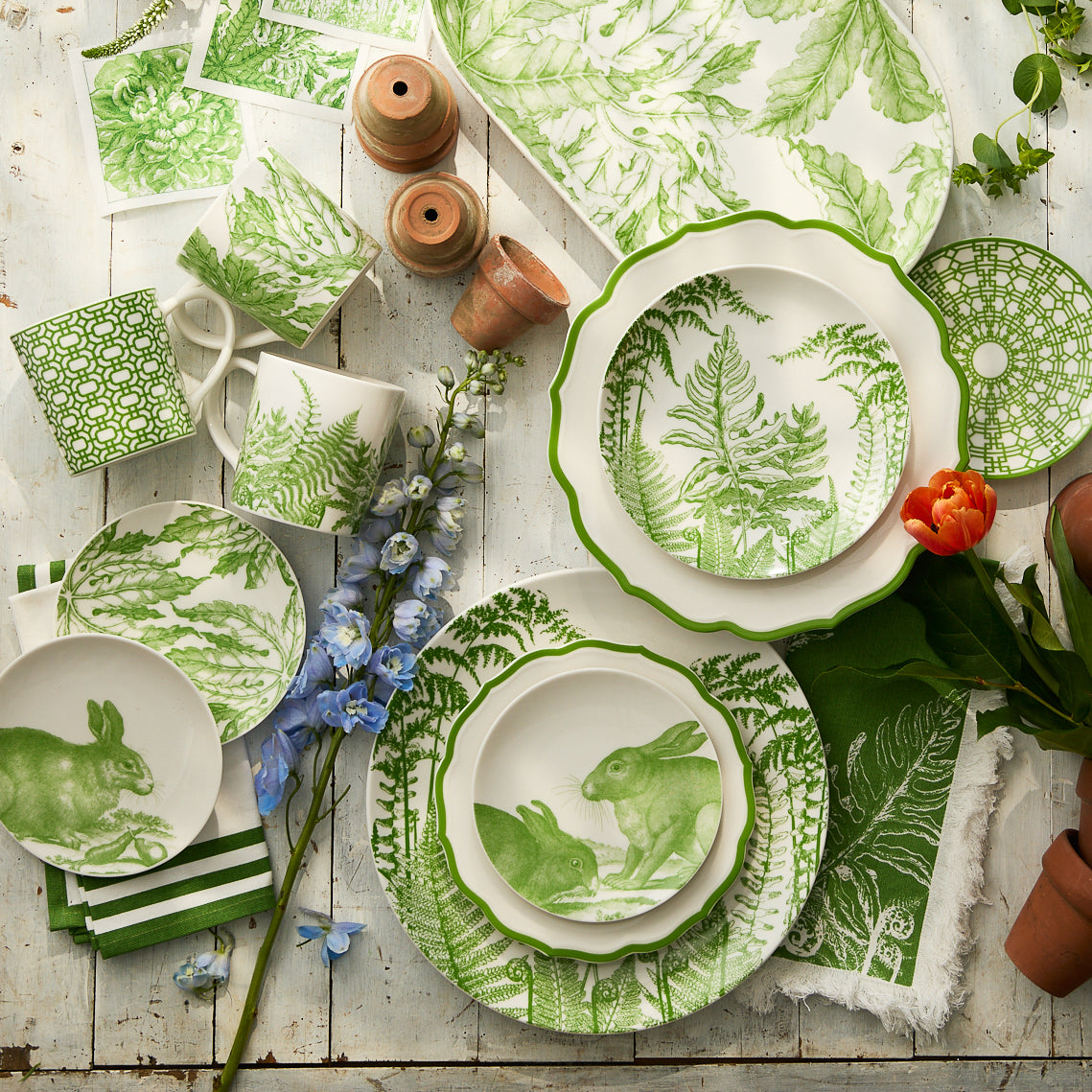 Assorted green and white tableware with botanical and rabbit motifs, flowers, Caskata Triple Stripe Verde Dinner Napkins (all machine washable), and small terra-cotta pots arranged on a rustic wooden surface.