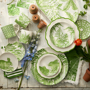 Assorted green and white tableware with botanical and rabbit motifs, flowers, Caskata Triple Stripe Verde Dinner Napkins (all machine washable), and small terra-cotta pots arranged on a rustic wooden surface.