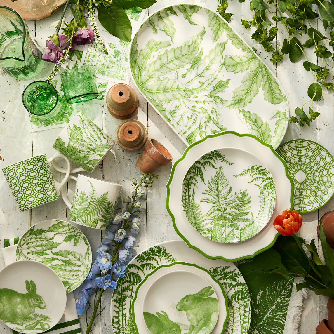 Caskata's Spring Verde Coupe Salad Plates, along with green and white dishes, rabbit-themed bowls and mugs, are displayed on a white wooden surface with green glassware and assorted flowers.