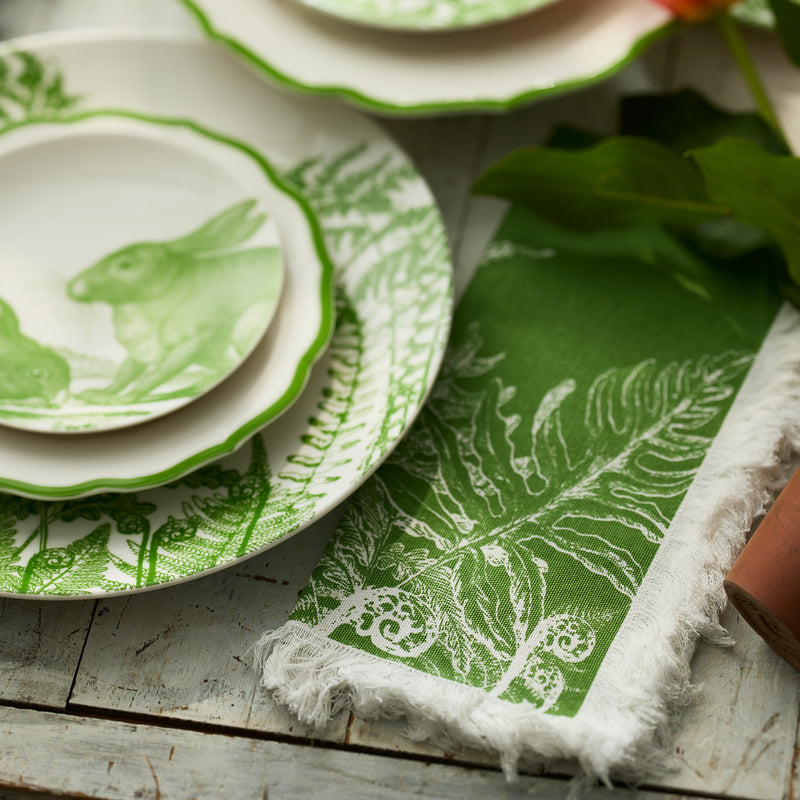 Green and white plates with rabbit and fern designs sit on a rustic wooden table beside Caskata's Spring Verde Dinner Napkins, featuring a fern pattern—ideal for fresh spring table decor.