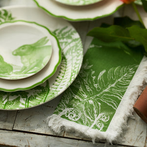 Green and white plates with rabbit and fern designs sit on a rustic wooden table beside Caskata's Spring Verde Dinner Napkins, featuring a fern pattern—ideal for fresh spring table decor.
