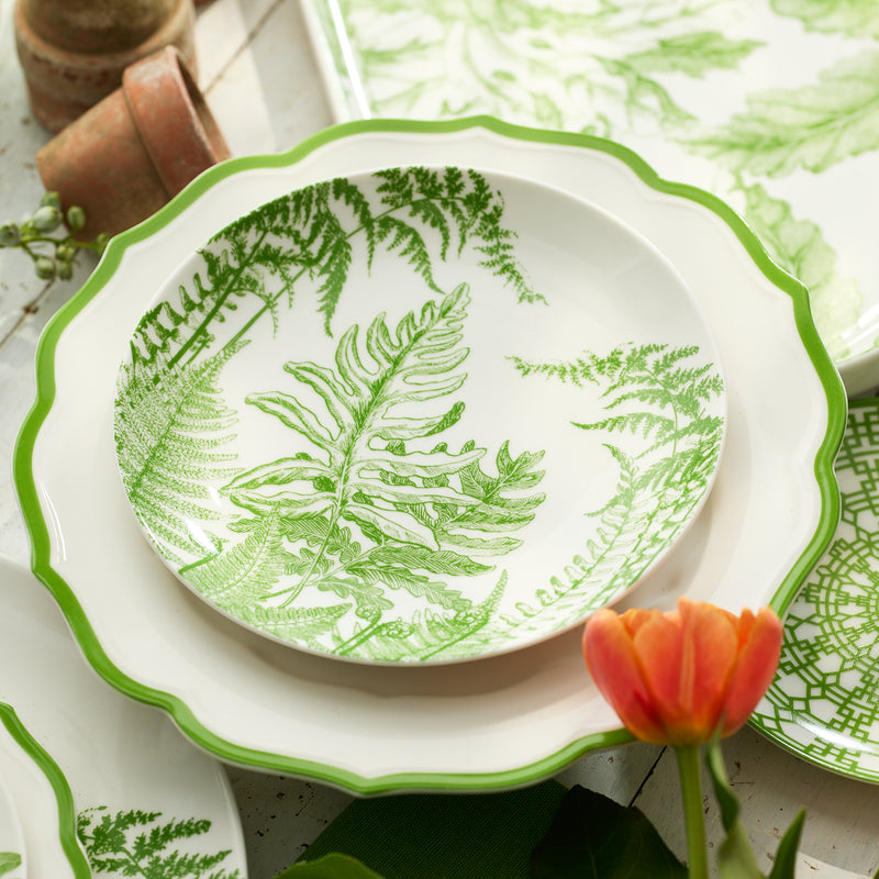 The Caskata Spring Verde Coupe Salad Plate features green fern leaf patterns and is stacked by a small orange tulip and plant pots, adding fresh botanical charm to your table.