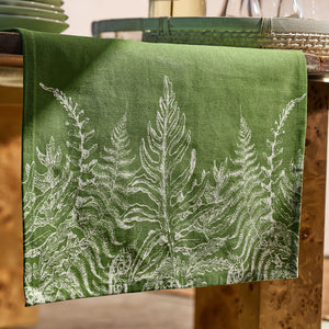 The Caskata Spring Verde Table Runner features a white botanical fern pattern on cotton, draping elegantly over a wooden table. Paired with glassware and a green tray, it creates charming cottage-core décor for any setting.