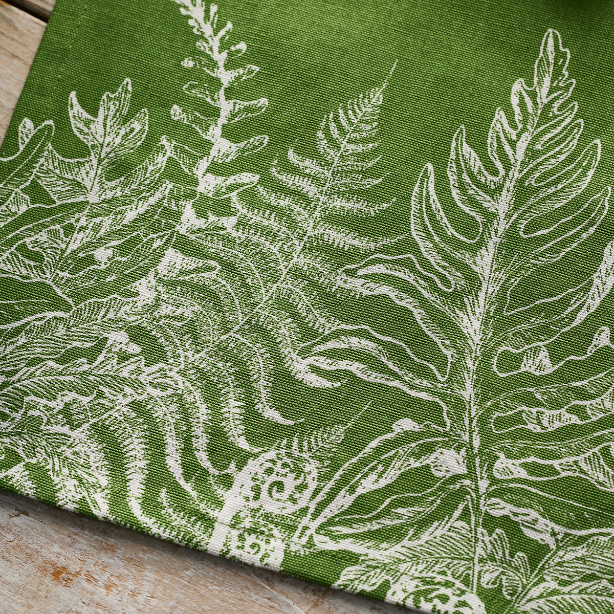 The Caskata Spring Verde Table Runner features green fabric with a white fern and leaf pattern, adding a cottage-core charm to your table décor.