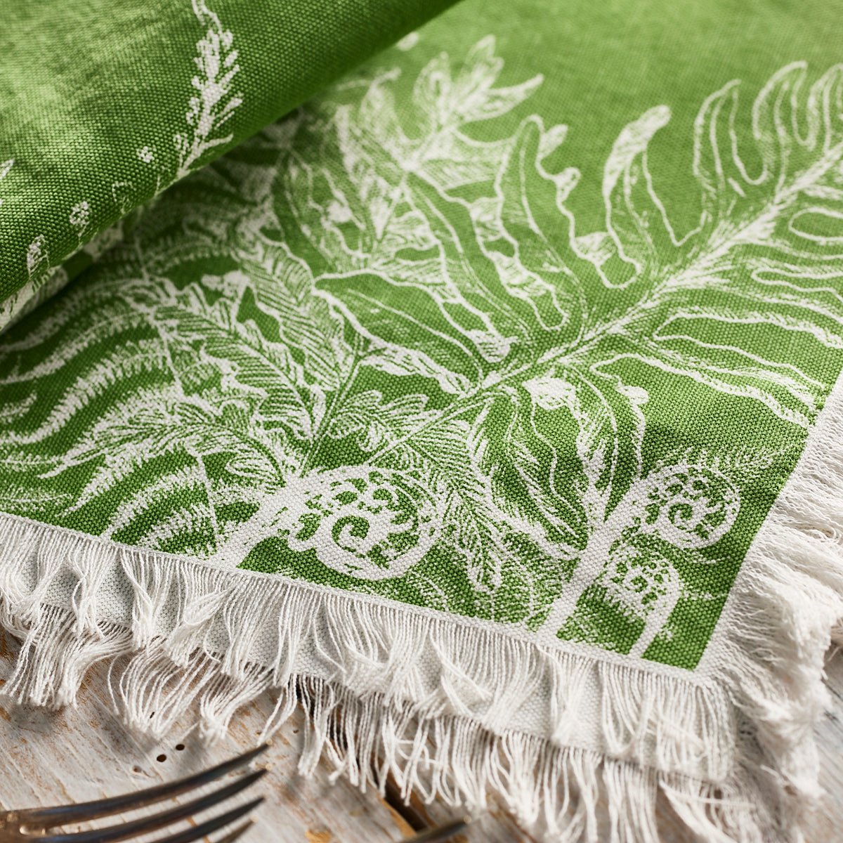 A green cotton napkin with white botanical patterns and fringed edges from Caskata’s Spring Verde Dinner Napkins rests on a rustic wooden surface beside metal forks, adding fresh seasonal charm to the table.