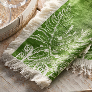 A Caskata Spring Verde Dinner Napkin with white botanical prints and fringed edges lies on a light wood surface beside a woven tray with glassware.