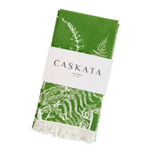 A folded green fern napkin with white botanical designs and fringe, wrapped in a band labeled 