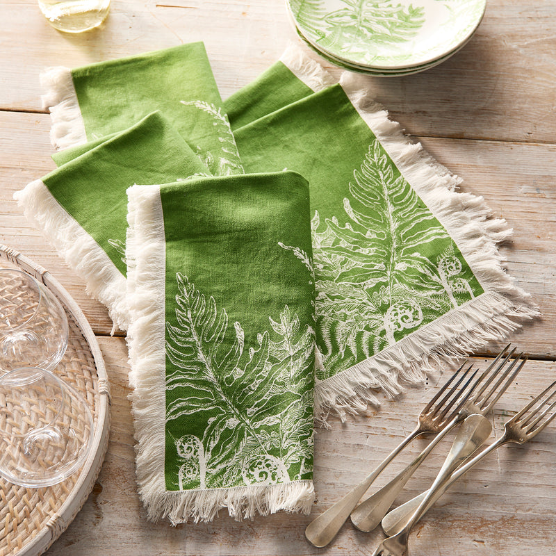 Caskata’s Spring Verde Dinner Napkins—green cotton with white fringe and a fern design—bring fresh charm to your tablescape, shown here next to silverware, glassware, and a matching plate on a rustic wooden table.