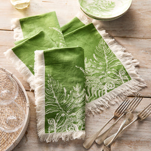 Caskata’s Spring Verde Dinner Napkins—green cotton with white fringe and a fern design—bring fresh charm to your tablescape, shown here next to silverware, glassware, and a matching plate on a rustic wooden table.