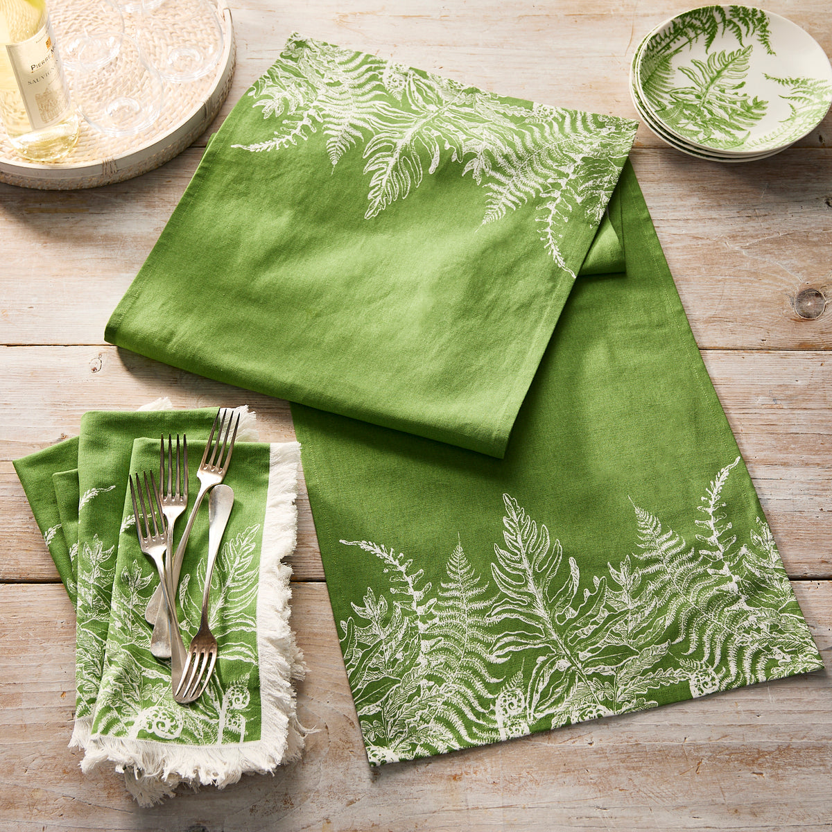 Caskata’s Spring Verde Dinner Napkins, featuring green linens with white fern patterns, are set on a wooden table beside a small matching bowl and silver forks resting on one napkin.