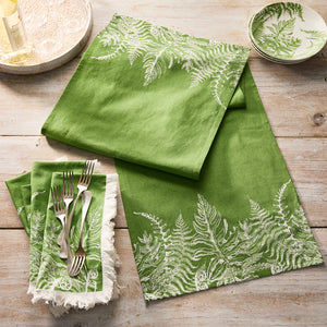 Caskata’s Spring Verde Dinner Napkins, featuring green linens with white fern patterns, are set on a wooden table beside a small matching bowl and silver forks resting on one napkin.