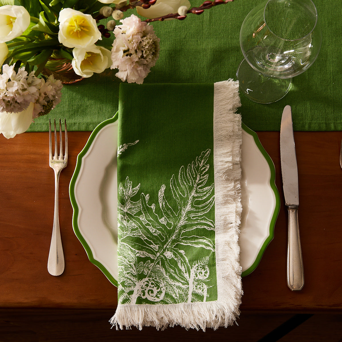 A white plate with a green rim is set with a fork, knife, and glass. Caskata Spring Verde Dinner Napkins with white fern print and fringe top the plate, while flowers and a green tablecloth complete the spring table decor.