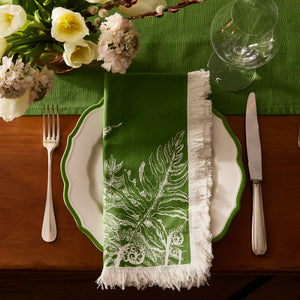 A white plate with a green rim is set with a fork, knife, and glass. Caskata Spring Verde Dinner Napkins with white fern print and fringe top the plate, while flowers and a green tablecloth complete the spring table decor.