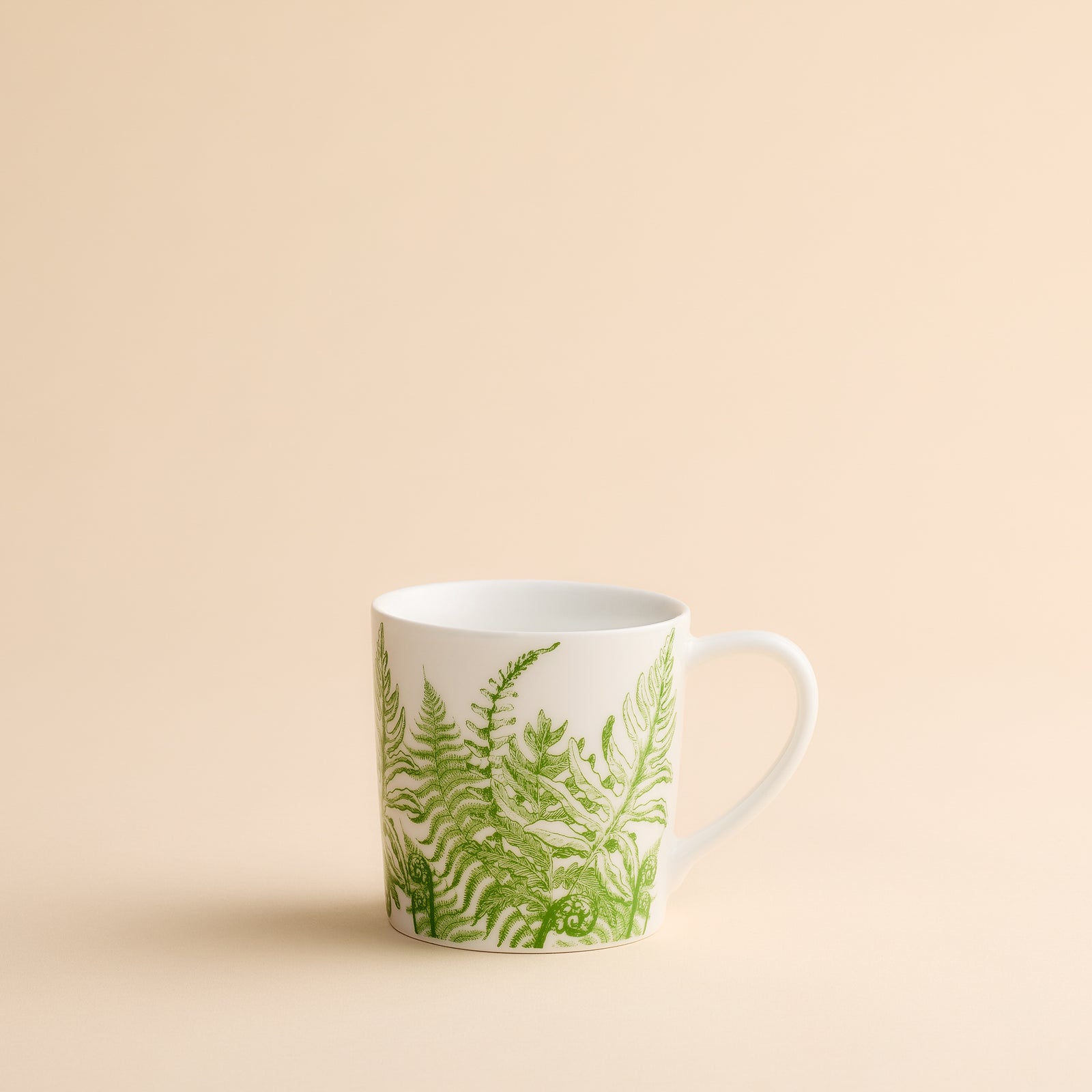 Elegant Caskata Spring Verde Mug crafted from white porcelain and decorated with green fern leaf motifs, set against a neutral beige background—a perfect botanical addition to your mug collection.