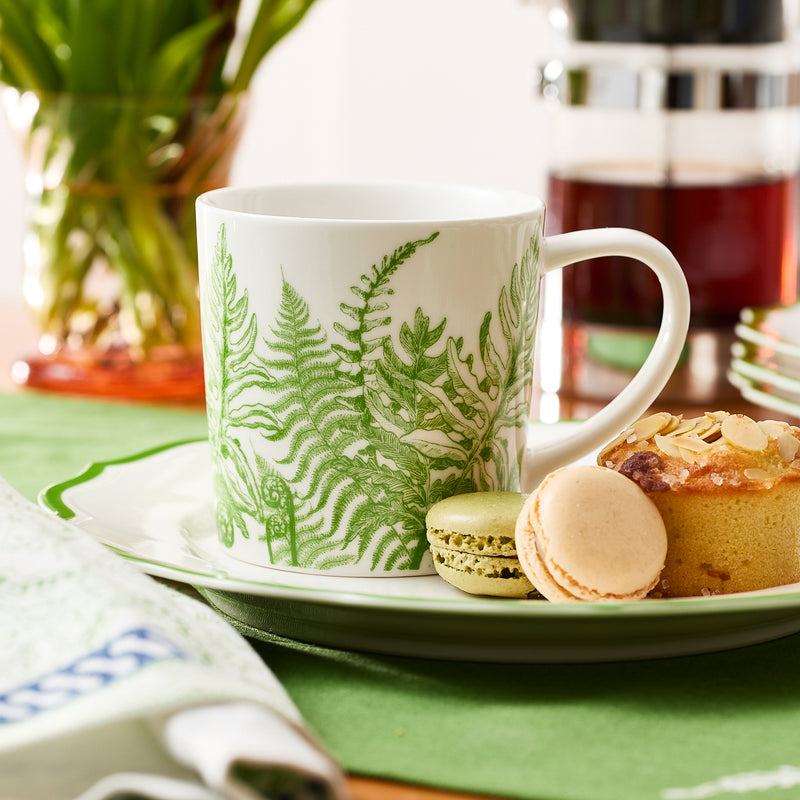 A Caskata Spring Verde Mug, decorated with green fern designs, rests on a plate alongside two macarons and a pastry, while a French press and flowers add brightness to the scene.